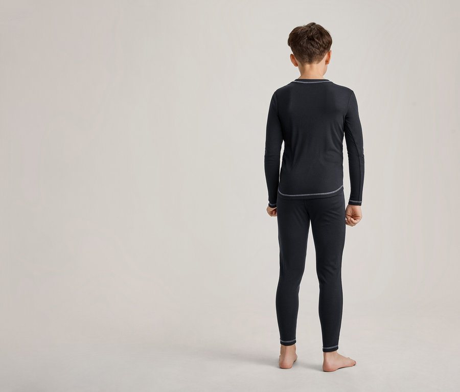 Ein junger Junge steht mit dem Rücken zum Betrachter und trägt ein schwarzes Langarmshirt und schwarze Leggings mit sichtbaren grauen Nähten. Er ist barfuß.