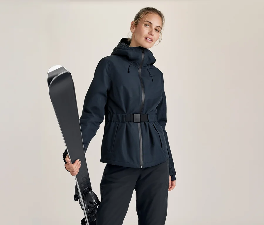 Frau in dunkelblauer, taillierter Skijacke und dunkler Hose, die ein Paar schwarze Skier über der linken Schulter hält.