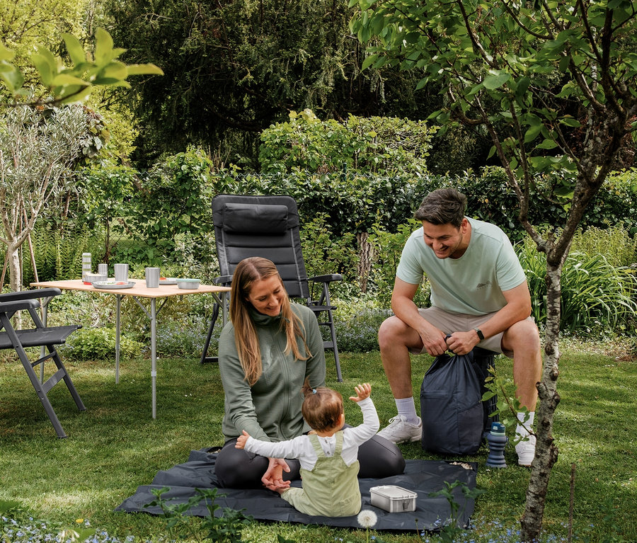 Eine Frau in grüner Funktionsjacke und ein Baby sitzen auf einer dunkelgrauen Mini-Picknickdecke im Garten, daneben sitzt ein lächelnder Mann.