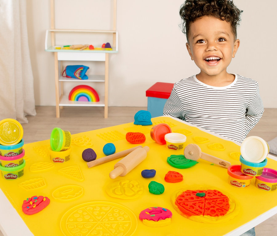 Lächelnder Junge am roba x Play-Doh Sitzgruppen-Set mit gelber Matte, Play-Doh, Nudelholz und Förmchen.