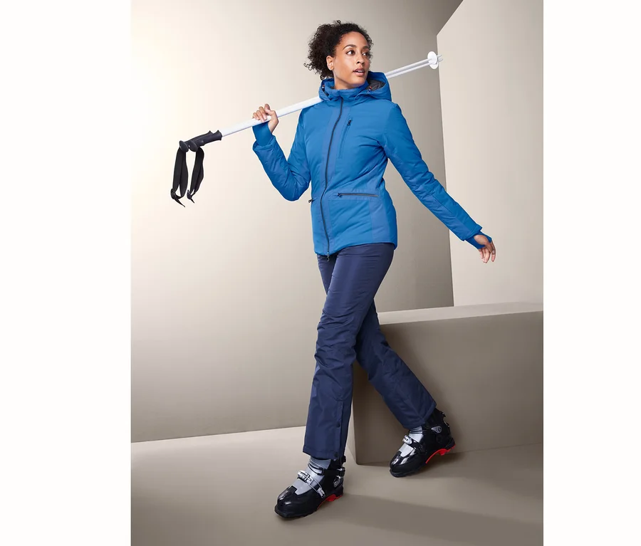 Frau geht mit Skistöcken über der Schulter und trägt eine Softshell-Skijacke.