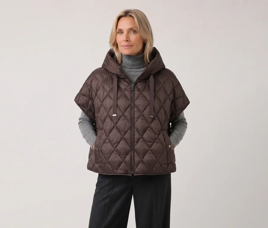 Eine blonde Frau trägt eine braune Barbara Lebek Steppweste mit Kapuze und kurzen Ärmeln über einem grauen Rollkragenpullover und einer dunklen Hose.