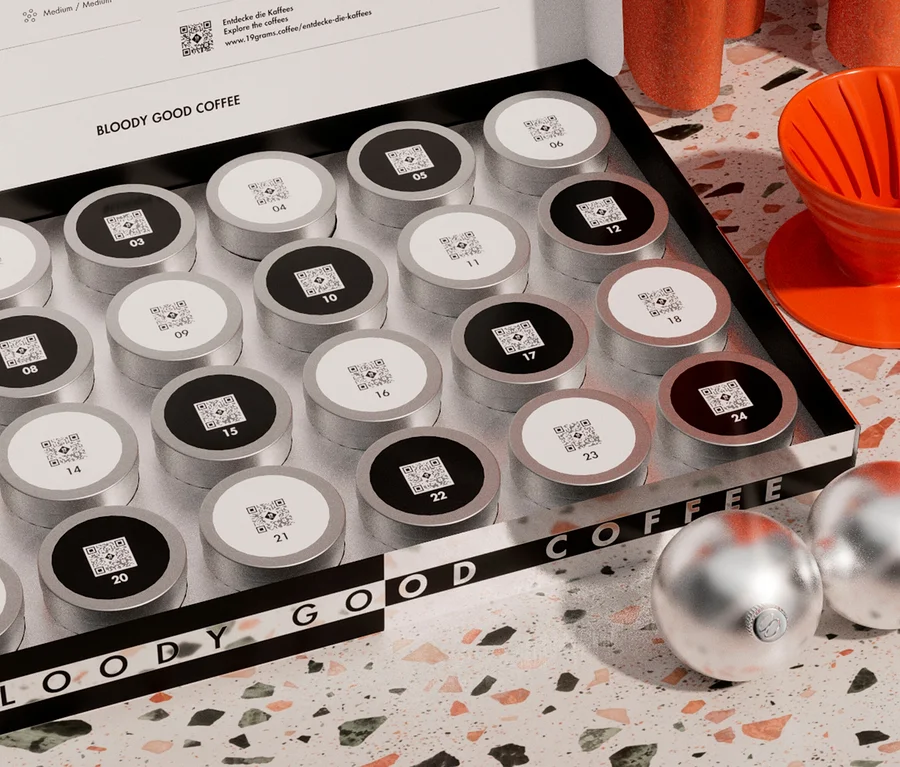 Eine offene Box mit 24 silbernen Kaffeekapseln mit schwarzen und weißen Deckeln, nummeriert und mit QR-Codes versehen. Rechts daneben ein orangefarbener Kaffeefilter mit Untertasse und im Vordergrund zwei silberne Christbaumkugeln auf einer Terrazzo-Oberfläche.