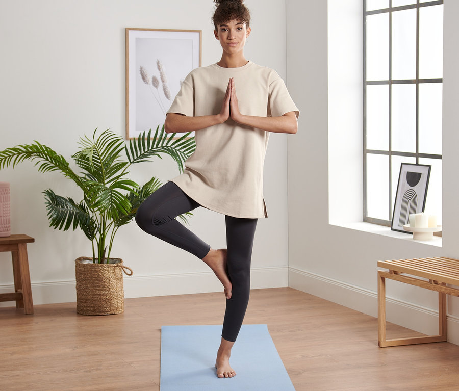 Frau steht in Baumpose auf einer blauen Yogamatte und trägt ein beiges Oversized-Sportshirt.