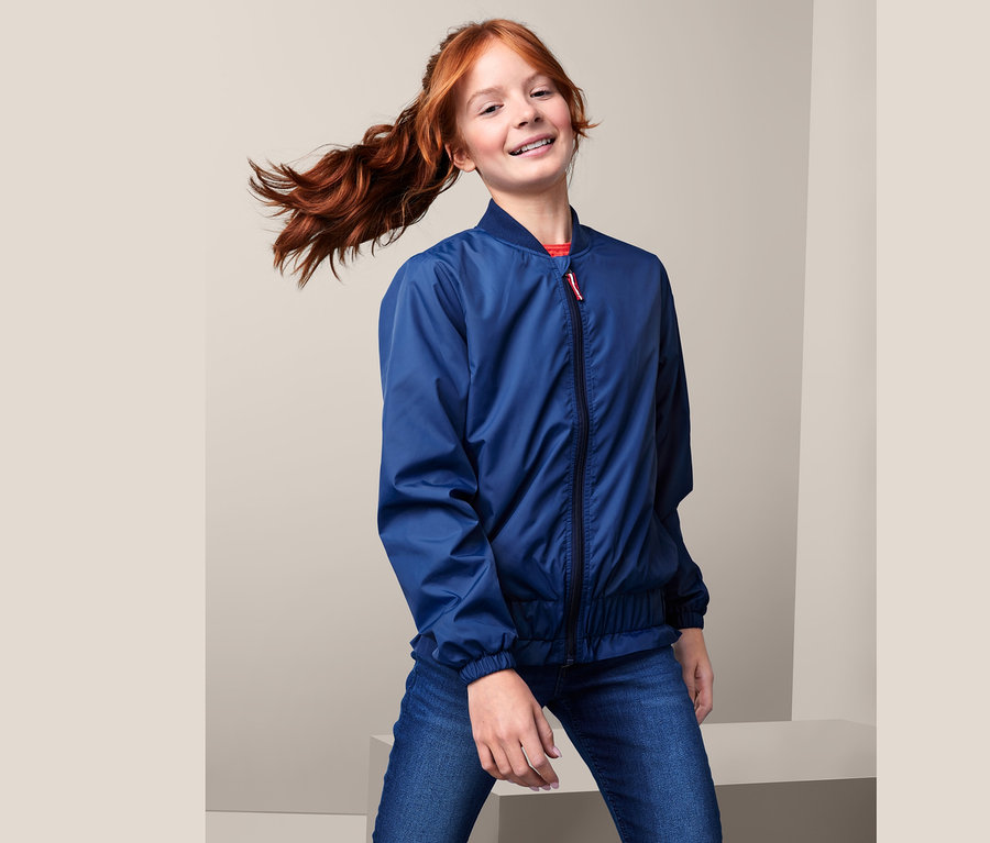Ein Mädchen mit einem roten Pferdeschwanz posiert in einer blauen, recycelten Bomberjacke und Jeans.