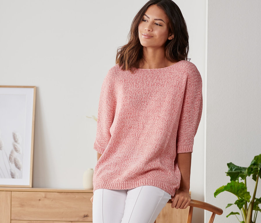 Eine Frau, die sich an einen Stuhl lehnt, trägt einen sommerlichen Strickpullover aus Bändchengarn, weiße Hosen und Turnschuhe.