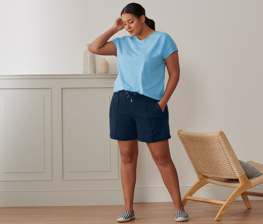 Eine Frau steht in einem Raum und trägt ein hellblaues Shirt aus Leinenmix, Sweatshorts und Espadrilles.