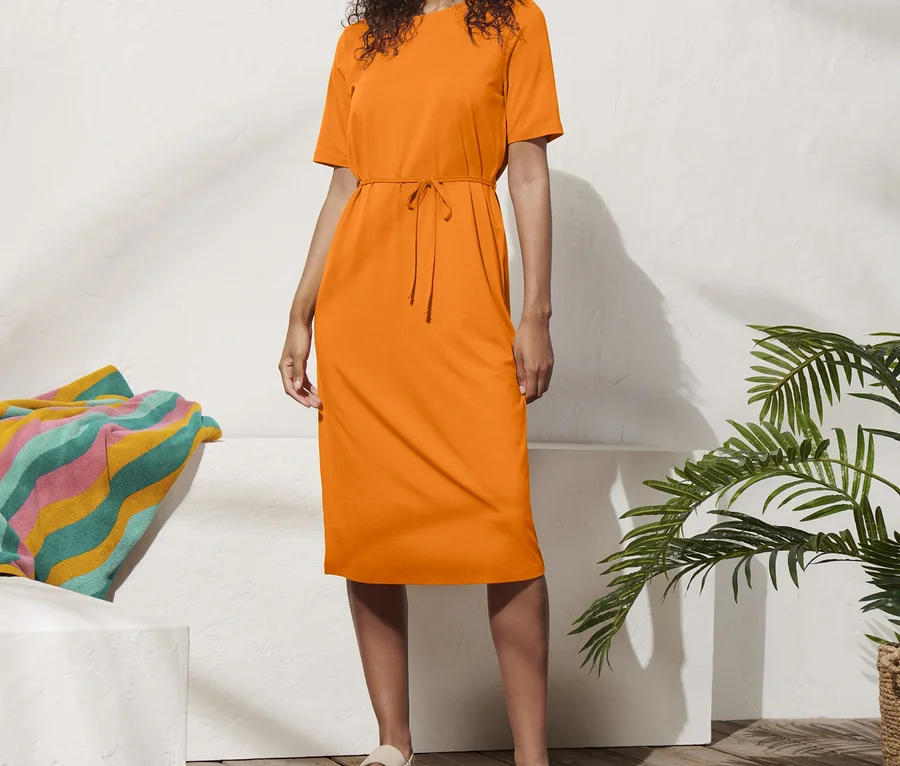 Frau steht in einem orangefarbenen Jerseykleid mit Taillengürtel und Hut.