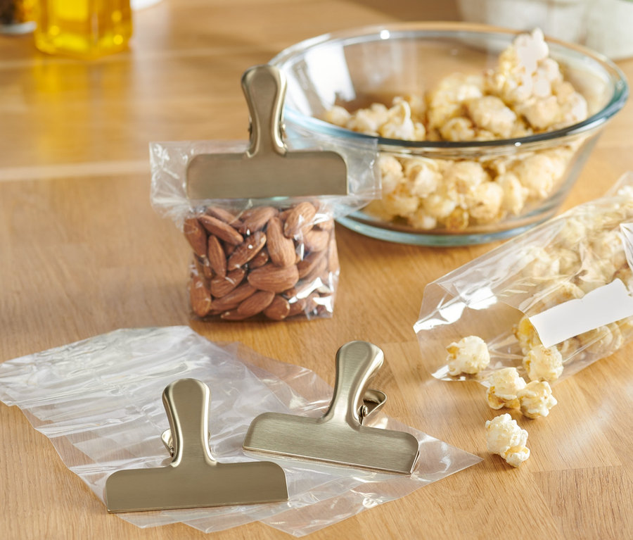 Auf einem Holztisch liegen 3 Edelstahl-Aromaclips, zwei davon auf leeren Tüten und einer auf einer Tüte mit Mandeln.