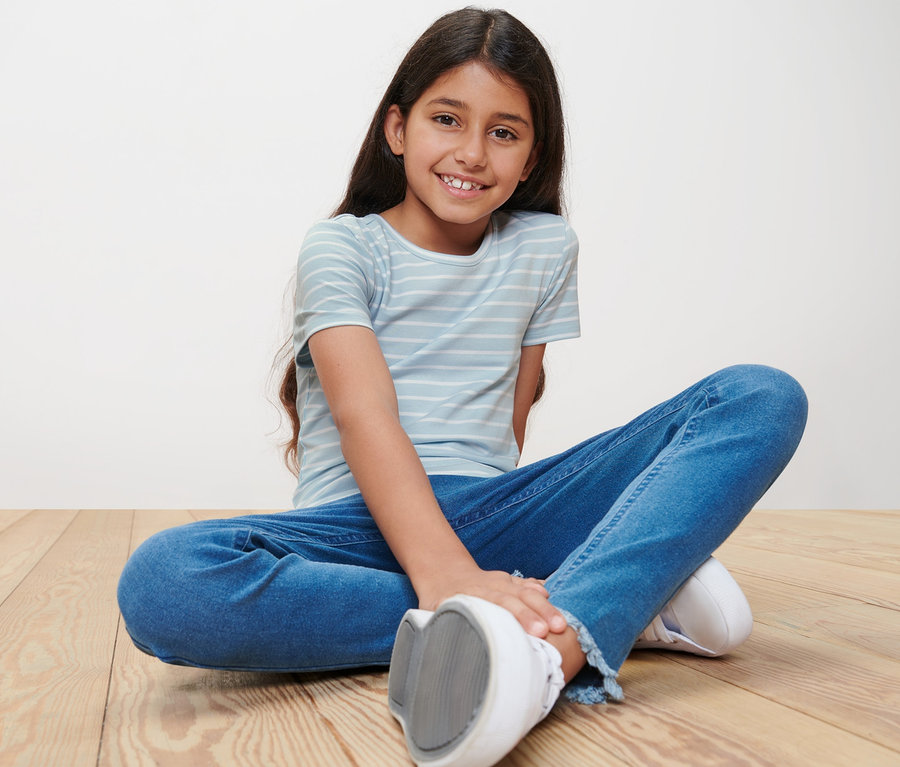 Ein lächelndes Mädchen sitzt in Jeans und einem gestreiften T-Shirt auf einem Holzboden. Sie trägt eines von 3 Kinder-T-Shirts.