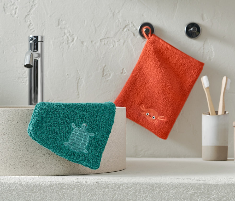 Ein grüner Waschlappen mit einer Schildkröte liegt auf einem Waschbecken, ein orangefarbener Waschlappen mit einer Krabbe hängt an der Wand.