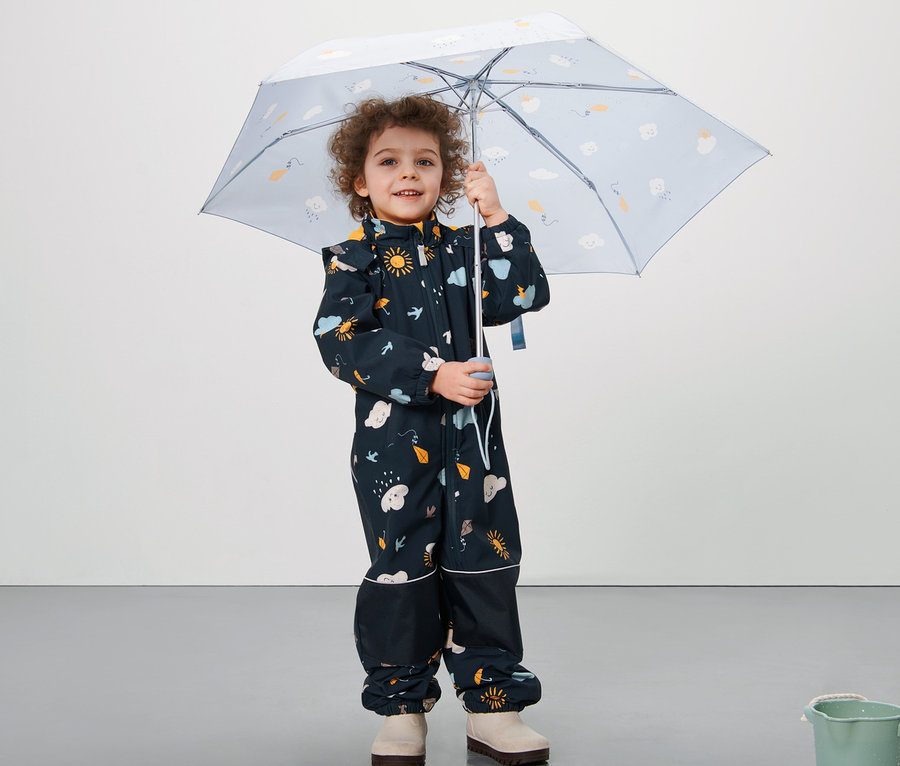 Junge hält einen Schirm und trägt einen blauen Kleinkind-Softshell-Einteiler mit Alloverprint.