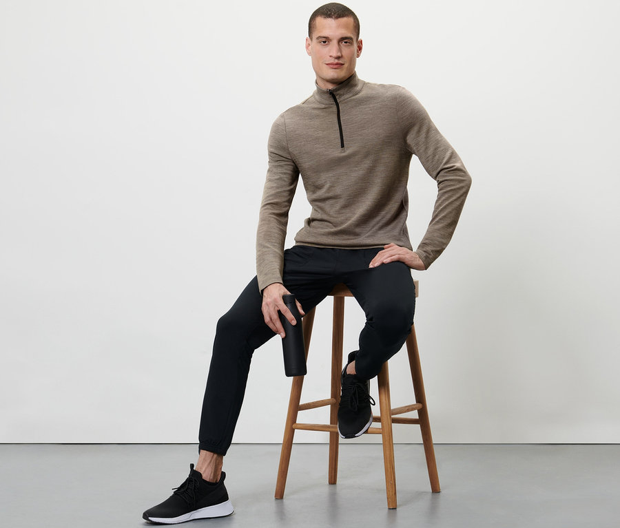 Mann sitzt auf einem Hocker und hält eine schwarze Flasche. Er trägt ein Shirt mit RWS-zertifizierter Wolle, schwarze Hosen und Turnschuhe.