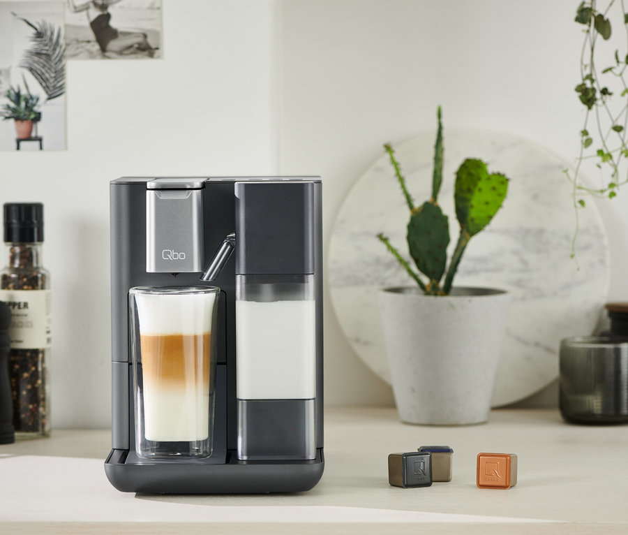 Graue Qbo MILK MASTER bereitet Getränk in Glas zu. Kapseln liegen auf Tisch.