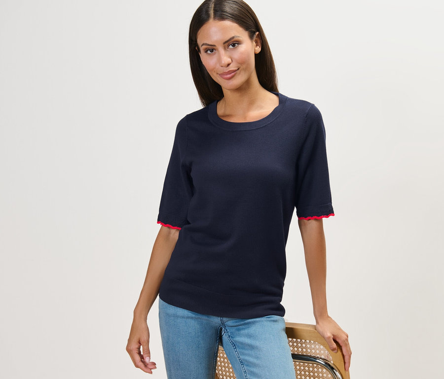 Frau posiert auf einem Stuhl und trägt einen dunkelblauen Feinstrickpullover und Jeans.