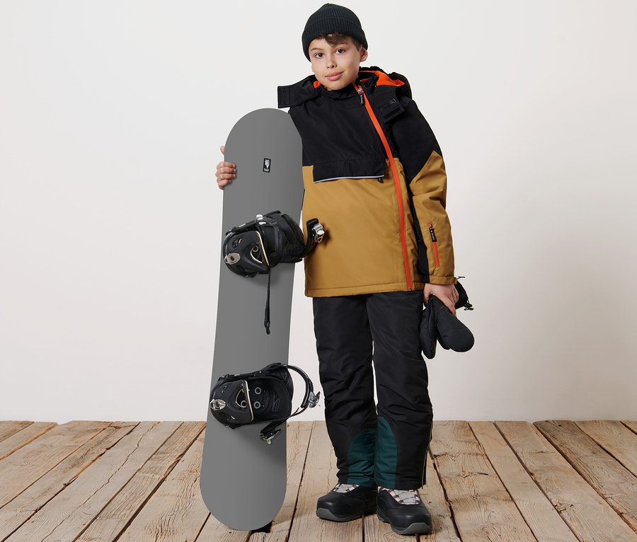 Junge hält Snowboard und trägt Kinder-Snowboardjacke.