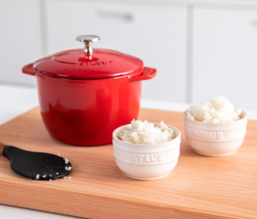 Ein roter STAUB Kochtopf »La Cocotte«, 16 cm, steht neben zwei Schalen Reis und einem schwarzen Löffel auf einem Holzbrett.