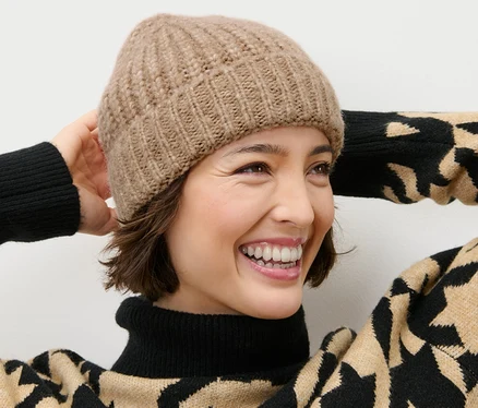 Lächelnde Frau trägt eine camel Strickmütze und einen Rollkragenpullover mit schwarzem und beigem Muster.