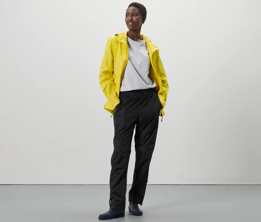 Eine Schwarze steht mit den Händen in den Taschen einer schwarzen High-Tech-Regenhose und einer gelben Regenjacke.
