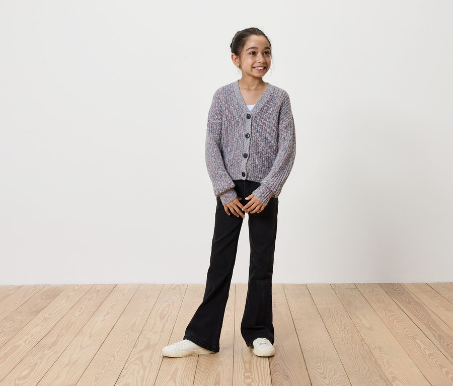 Lächelndes Mädchen in grauem Strickcardigan mit Knöpfen, schwarzer Schlaghose und weißen Turnschuhen steht auf einem Holzboden vor einer weißen Wand.