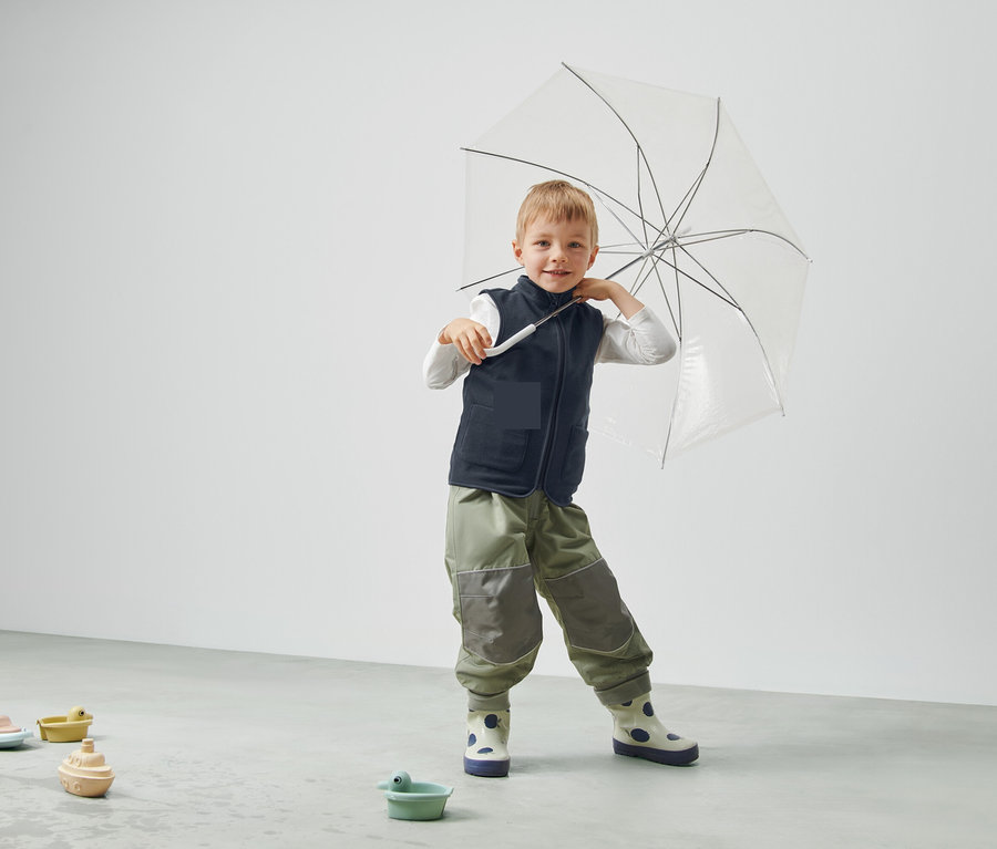 Junge hält einen durchsichtigen Regenschirm, trägt eine Weste und Hose und steht in einem Raum.