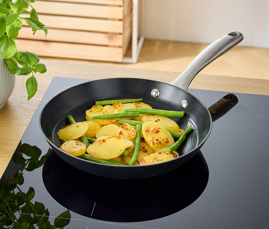 Eine Bratpfanne mit gebratenen Kartoffeln und grünen Bohnen liegt auf einem schwarzen Induktionskochfeld.