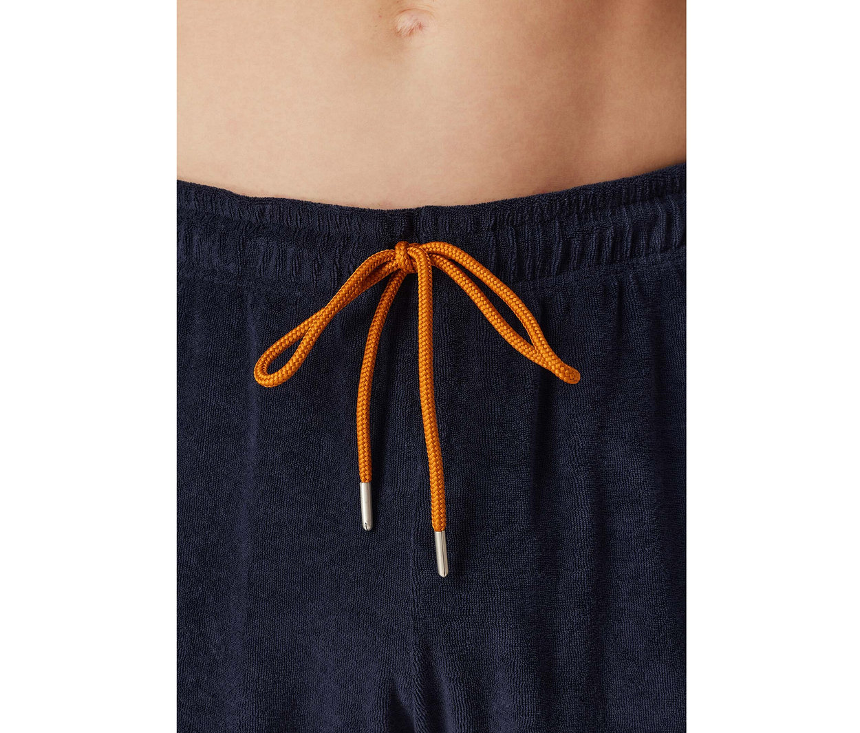Nahaufnahme des Bauches einer Person, die eine marineblaue SCHIESSER Frottee-Pyjamahose mit orangefarbenem Schleifenkordelzug trägt.