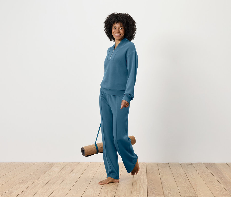 Lächelnde Frau mit Afro-Frisur geht barfuß und trägt eine Yogamatte, während sie ein blaues Trainingsanzug-Set trägt.