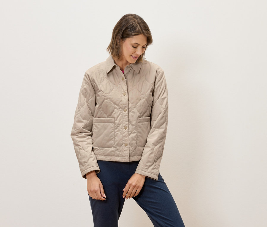 Eine Frau in einer gesteppten Jacke in Beige und blauer Hose steht vor einem weißen Hintergrund.