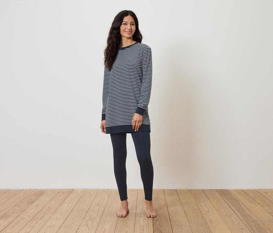 Eine barfuß stehende Frau auf einem Holzboden trägt ein gestreiftes Langarmshirt und dunkelblaue Leggings.