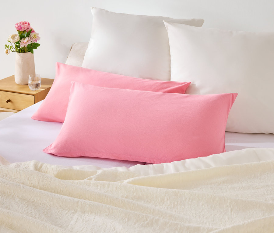 Auf einem Bett liegen zwei pinke und zwei weiße Kissen. Auf dem Nachttisch steht eine Vase mit Blumen.