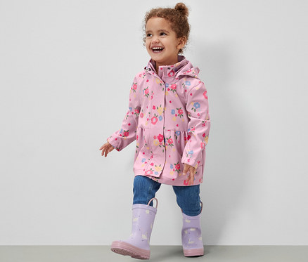 Lächelndes Mädchen in Kinder-Softshelljacke mit Blumenmuster, Jeans und lila Gummistiefeln geht auf grauem Boden vor einer weißen Wand.