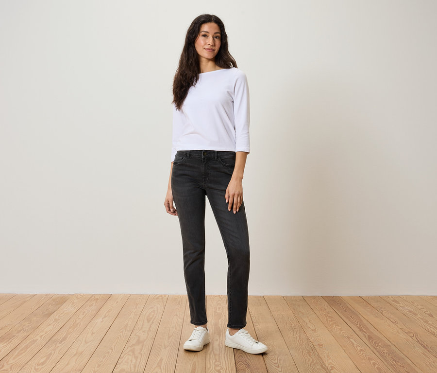 Eine Frau steht auf einem Holzboden und trägt ein weißes Dreiviertelarmshirt, graue Jeans und weiße Turnschuhe.