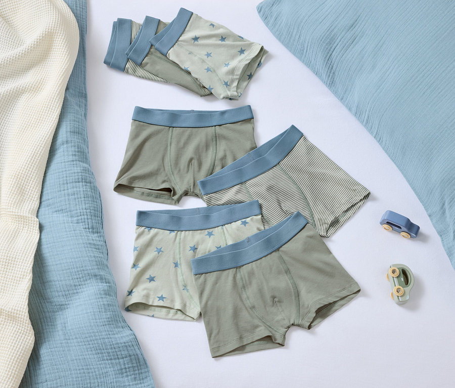 Fünf Boxershorts mit blauem Bund liegen auf einem weißen Hintergrund neben einem blauen Kissen und zwei kleinen Spielzeugen.