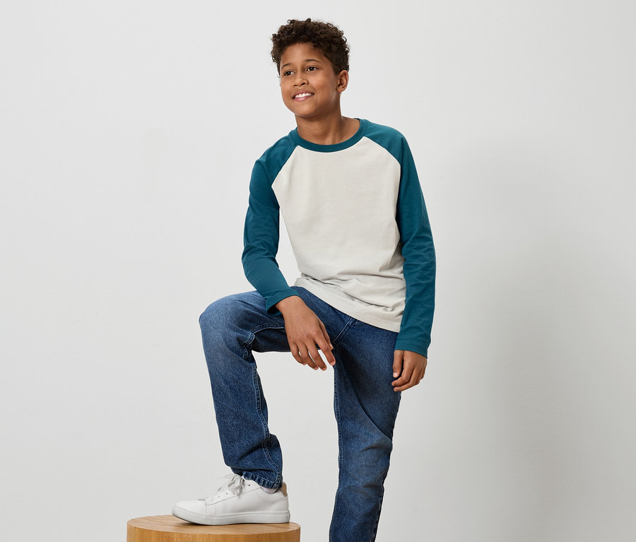 Ein junger, dunkelhaariger, lächelnder Junge posiert mit einem Bein auf einem Holzhocker, trägt blaue Jeans und weiße Turnschuhe.