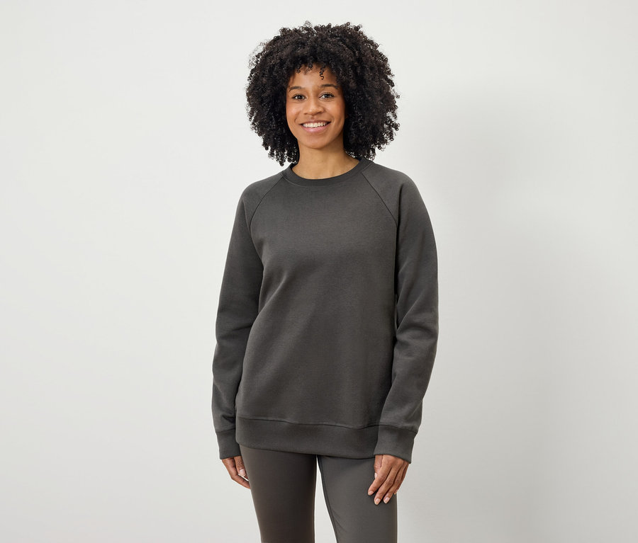 Eine Schwarze Frau mit lockigem Haar posiert vor einem hellen Hintergrund in einem grauen Sweatshirt und grauen Hosen.