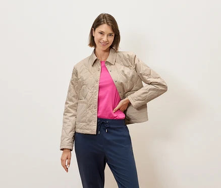 Eine Frau mit kurzem braunem Haar posiert vor einer cremefarbenen Wand in einer beige gesteppten Jacke, einem pinkfarbenen T-Shirt und blauen Hosen.