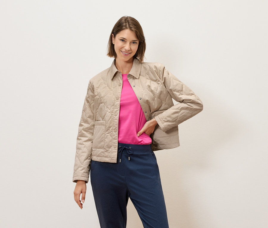 Eine Frau mit kurzem braunem Haar posiert vor einer cremefarbenen Wand in einer beige gesteppten Jacke, einem pinkfarbenen T-Shirt und blauen Hosen.