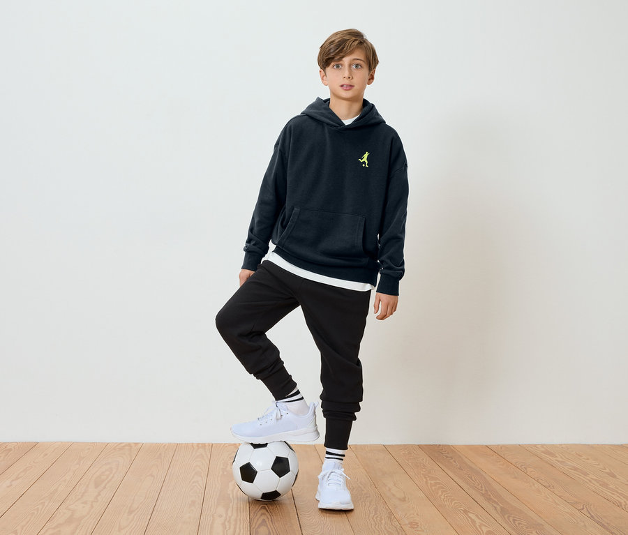Ein Junge steht mit einem Fuß auf einem Fußball, er trägt einen marineblauen Hoodie und schwarze Hosen.