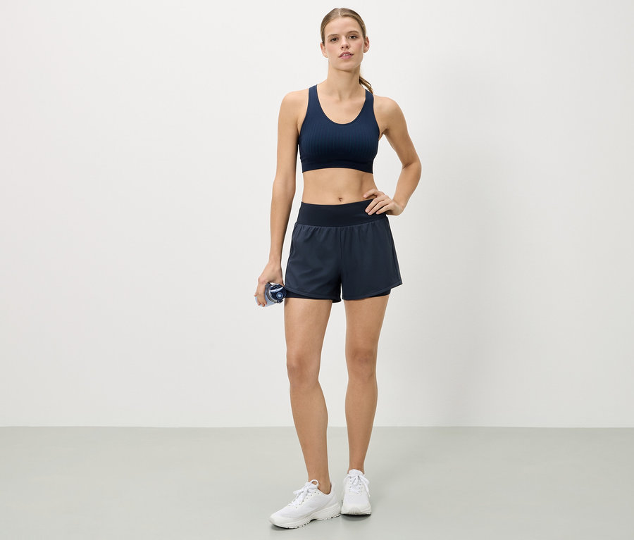 Frau steht mit der Hand auf der Hüfte, trägt einen marineblauen Sport-BH, Shorts und weiße Turnschuhe und hält eine Wasserflasche.