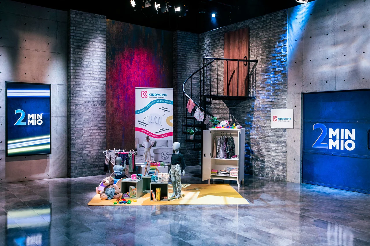 Auf einem Set mit Backsteinwänden stehen eine Wendeltreppe, ein Kleiderständer, Spielzeug und zwei Schaufensterpuppen.