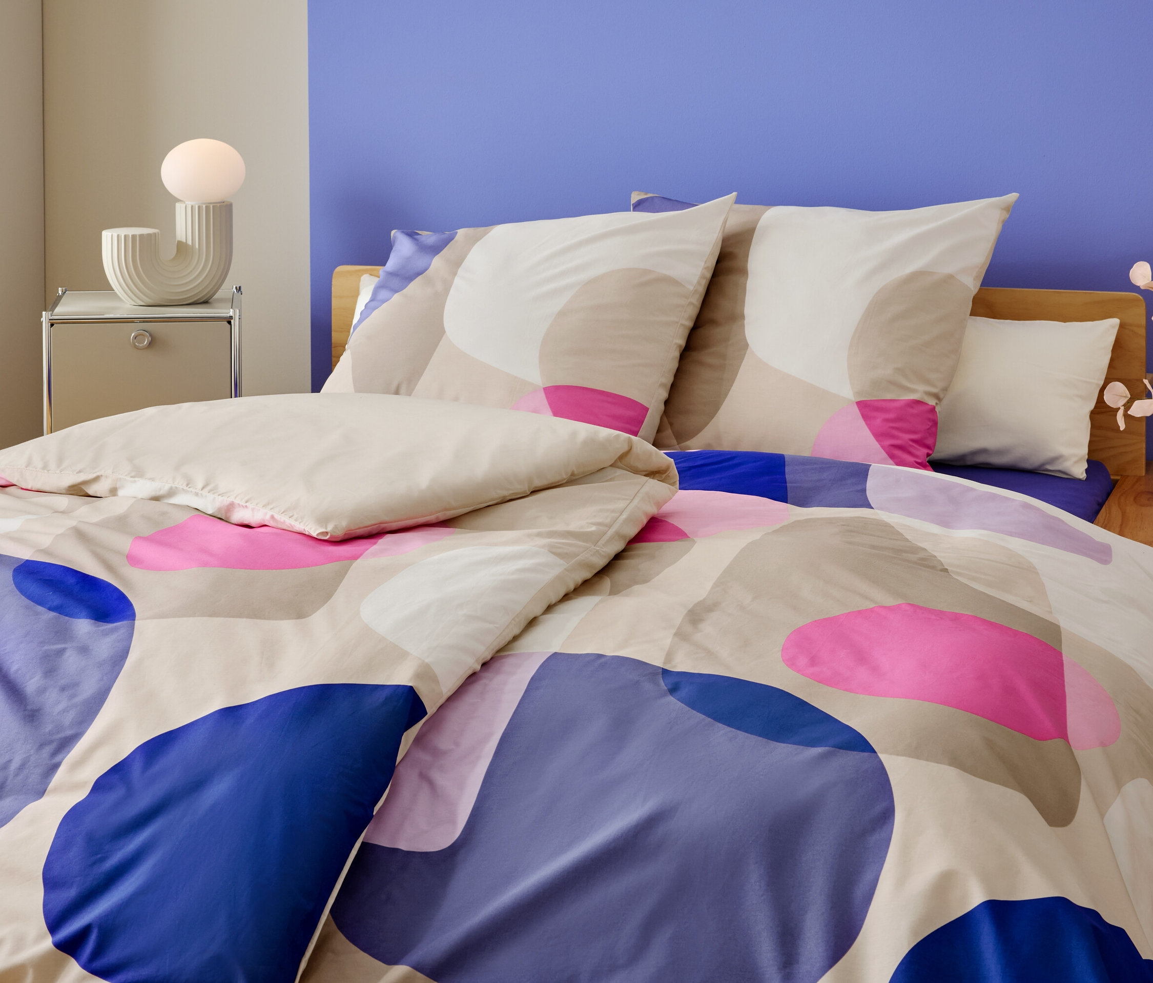 Nahaufnahme eines Bettes mit geometrischem Muster in Blau, Rosa und Beige. Auf dem Nachttisch steht eine Lampe und eine Vase mit Blumen.