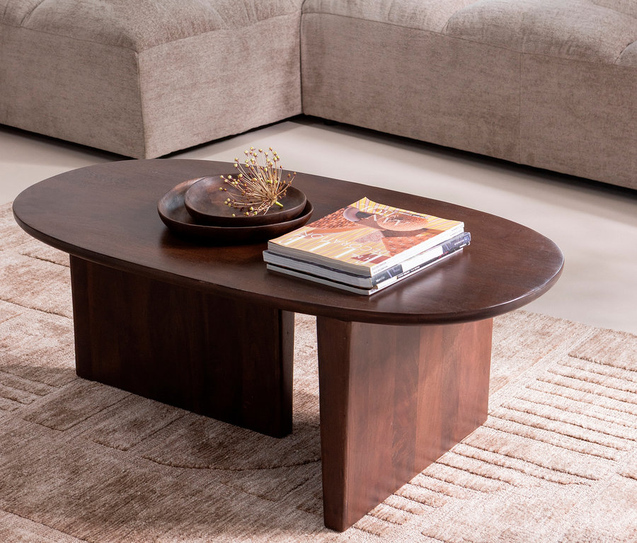 Ein ovaler, dunkelbrauner Couchtisch steht auf einem beigen Teppich. Auf dem Tisch liegen ein Magazin, eine Schale und eine kleine goldene Dekoration.