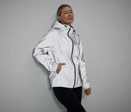 Frau mit Hand in Tasche, trägt reflektierende Jacke mit Kapuze und schwarzen Leggings vor grauem Hintergrund.