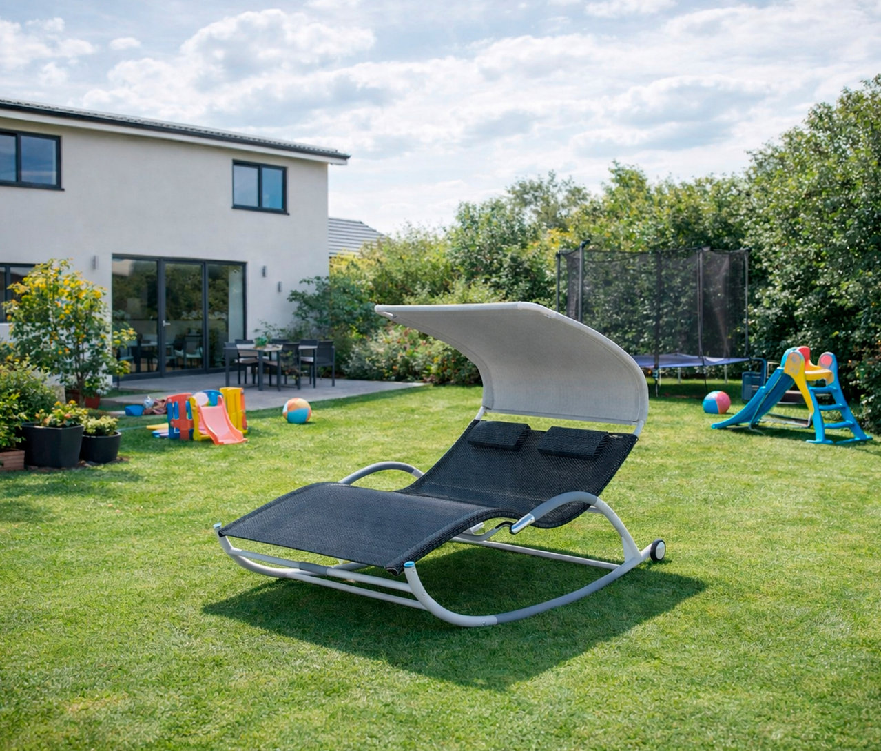 Im Garten steht eine Leco Doppelschaukelliege mit Rädern, im Hintergrund sind ein Haus, ein Trampolin und Spielsachen zu sehen.