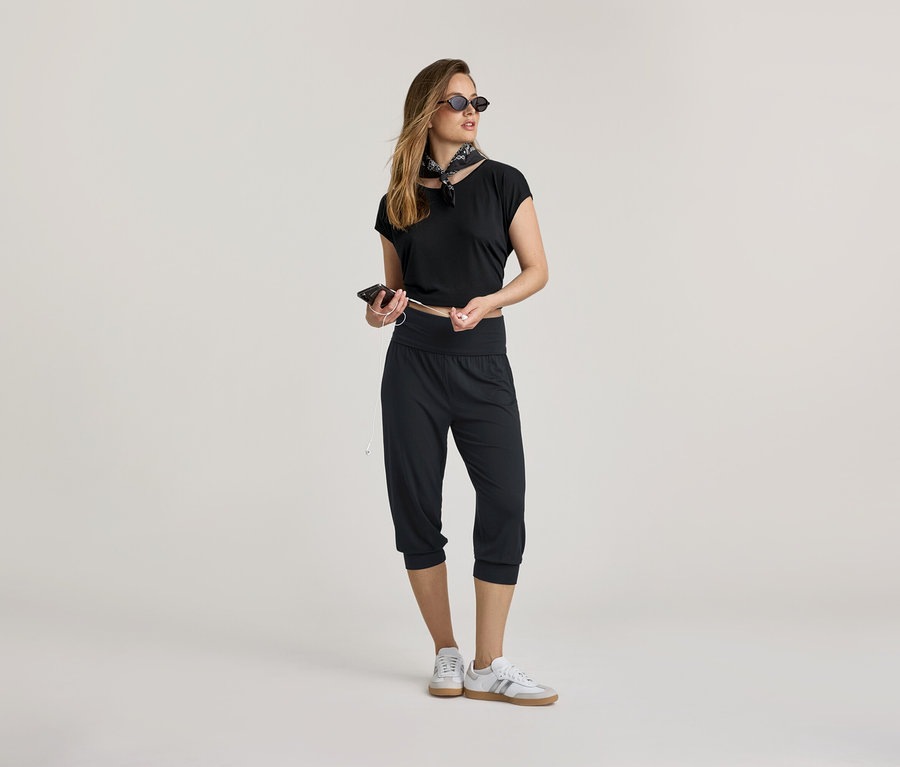 Frau in schwarzem Oberteil und 3/4-Yogahose mit Handy und Kopfhörern. Sie trägt ein Halstuch und Sneaker.