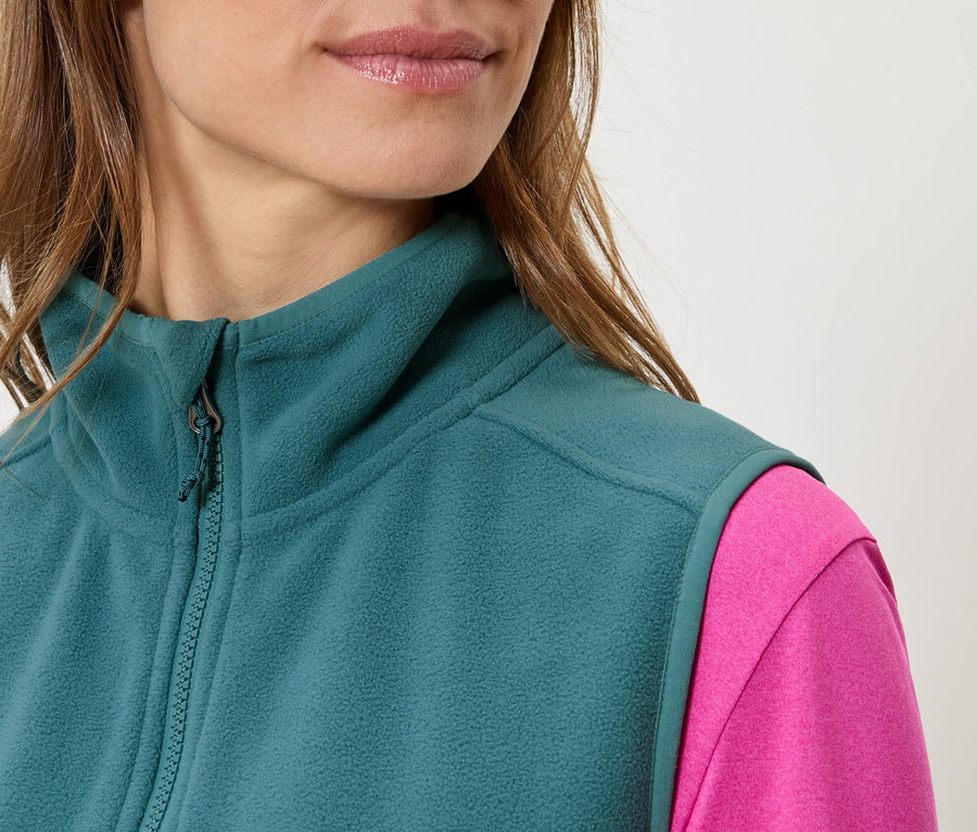 Nahaufnahme einer Frau, die eine petrolfarbene Fleeceweste über einem pinkfarbenen Oberteil trägt.