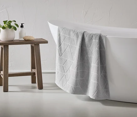 Ein graues Handtuch mit geometrischem Muster hängt über den Rand einer weißen Badewanne. Ein Holzhocker steht daneben, darauf eine Pflanze, Seife und ein Schwamm.