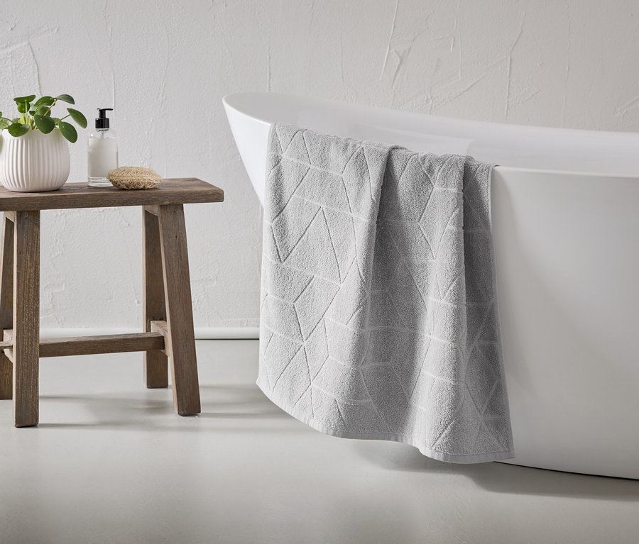 Ein graues Handtuch mit geometrischem Muster hängt über den Rand einer weißen Badewanne. Ein Holzhocker steht daneben, darauf eine Pflanze, Seife und ein Schwamm.