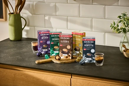 Fünf Packungen Eduscho Kaffeekapseln (Espresso Intenso, Lungo Cremoso, Lungo Elegante, Espresso Classico, Ristretto Perfetto) stehen auf einer Küchentheke mit zwei Tassen Kaffee und losen Kapseln.